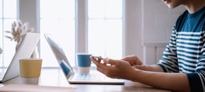 closeup of kitchen table, with person looking at phone next to computer and coffee cup